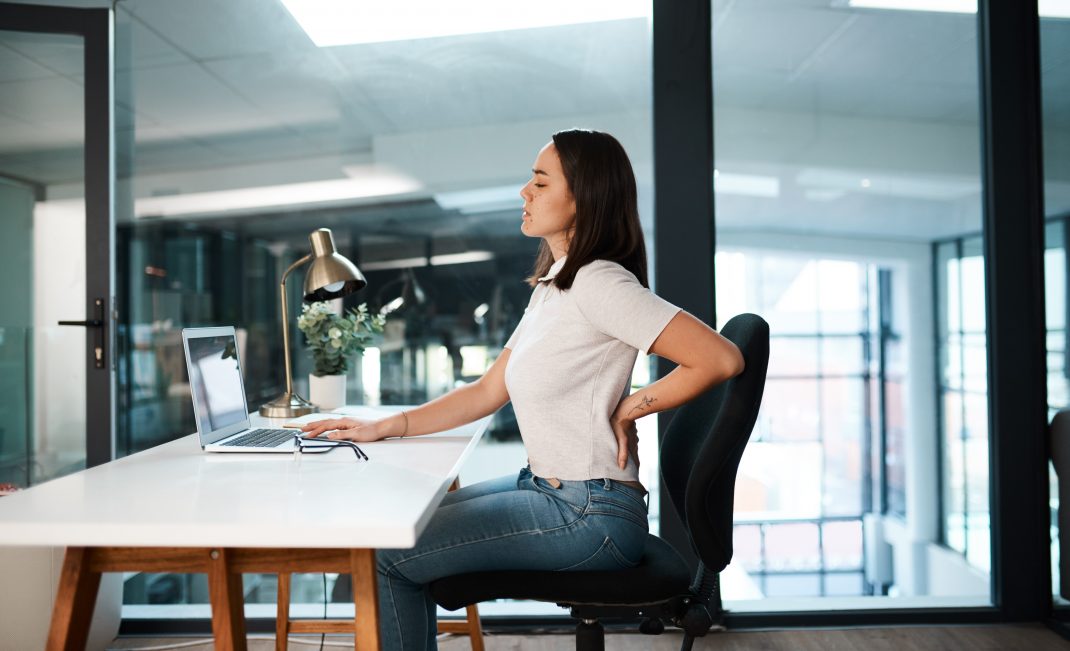 Woman sitting at work, holding her back, highlighting the adverse effects of desk jobs on health.