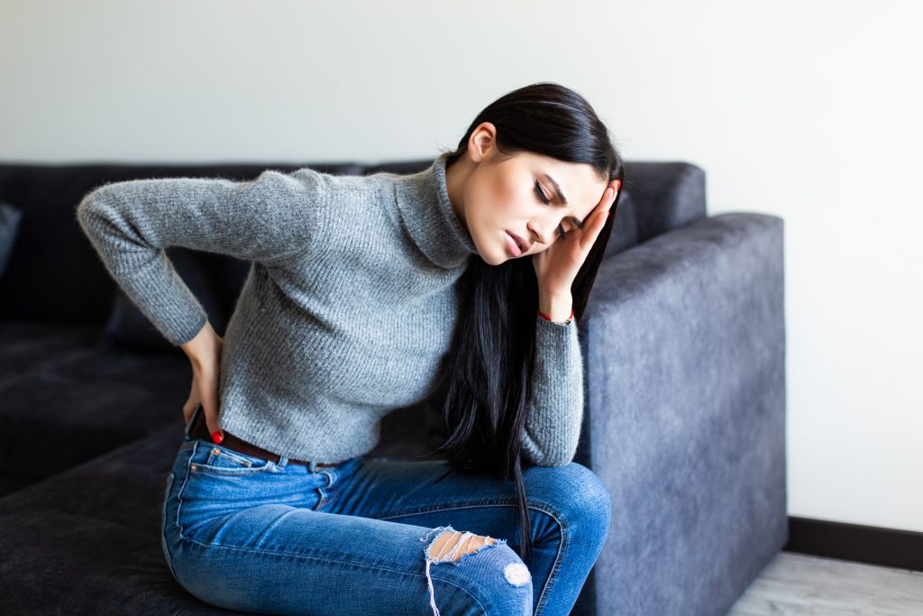 A woman sitting on a couch, holding her back in severe pain.