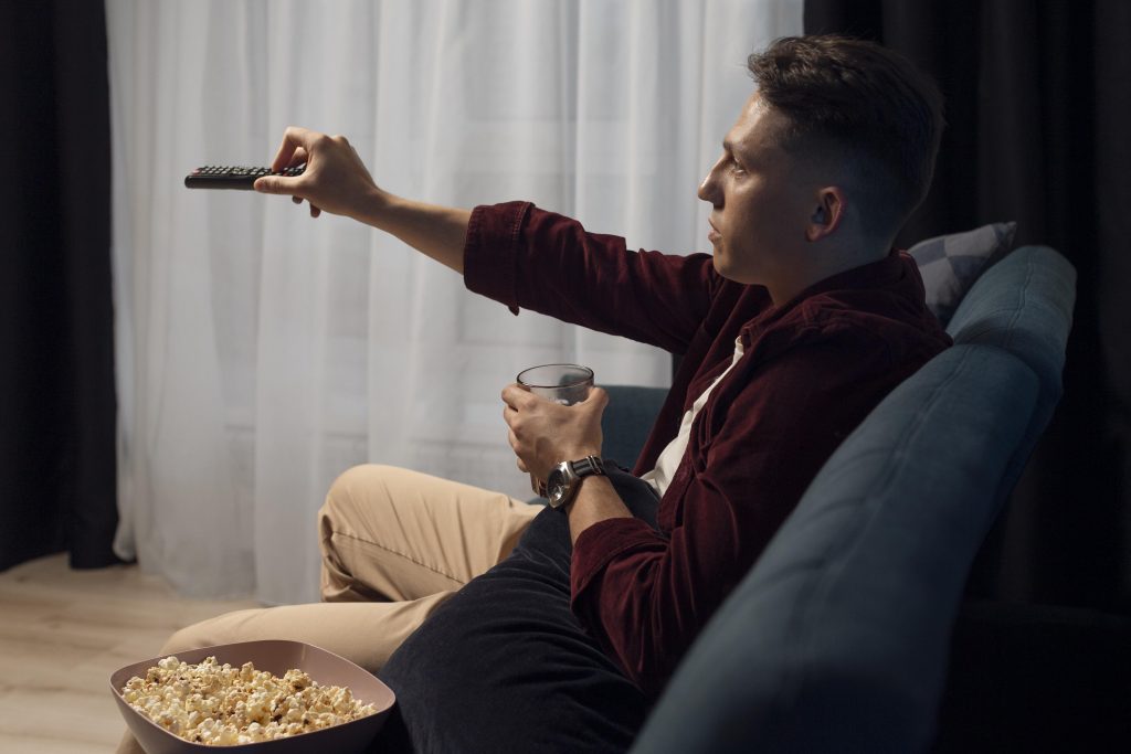 A man sitting on a sofa with a glass of drink in one hand and operating a remote in the other, watching TV.