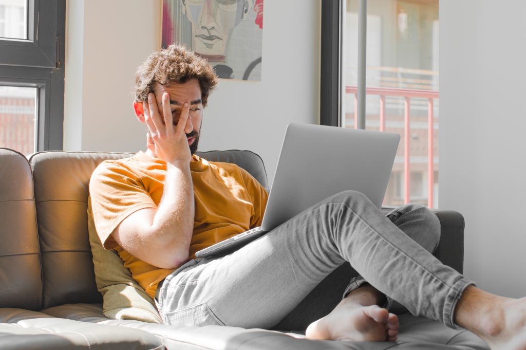 A man resting on a couch with tired eyes, binge-watching shows on his laptop.