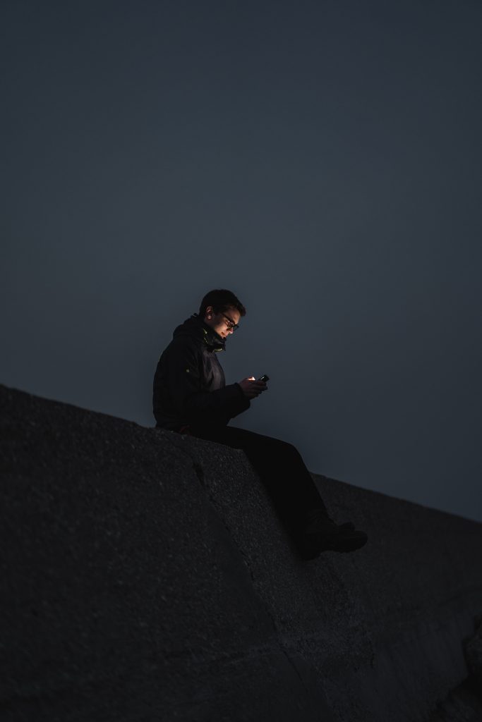 A man sitting alone outdoors in the dark, deeply engrossed in scrolling his phone.