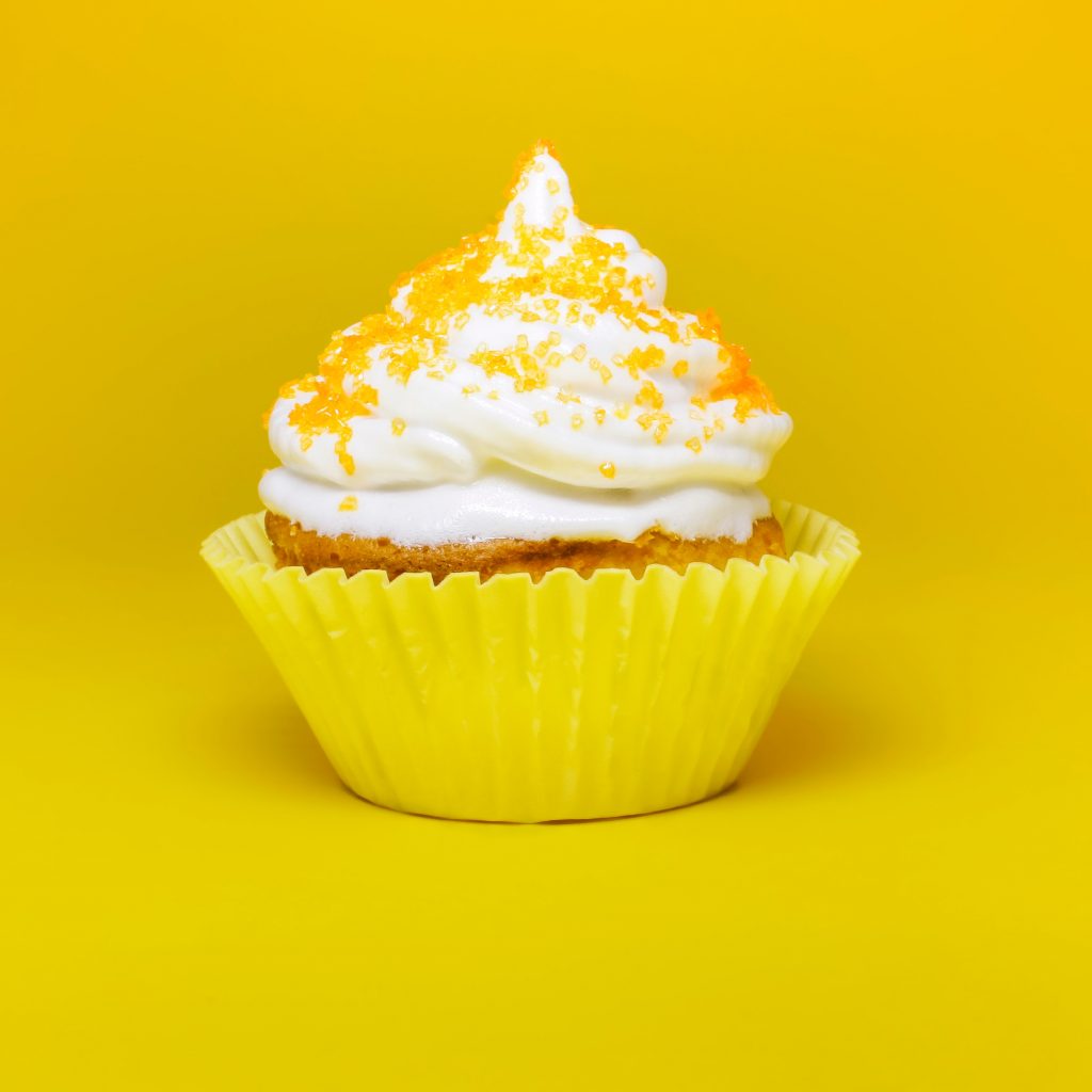 A photograph of a delicious-looking cupcake with a yellow wrapper, topped with yellow candies, elegantly displayed against a yellow background.