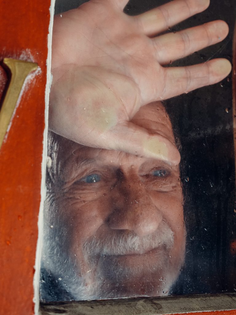 An elderly man standing by a window, smiling and lost in thought.