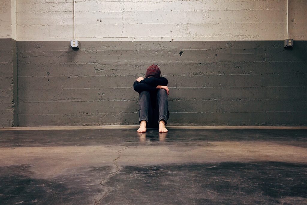 A person experiencing depression sitting alone on the floor in a large room, with their head bowed and hidden between their legs.