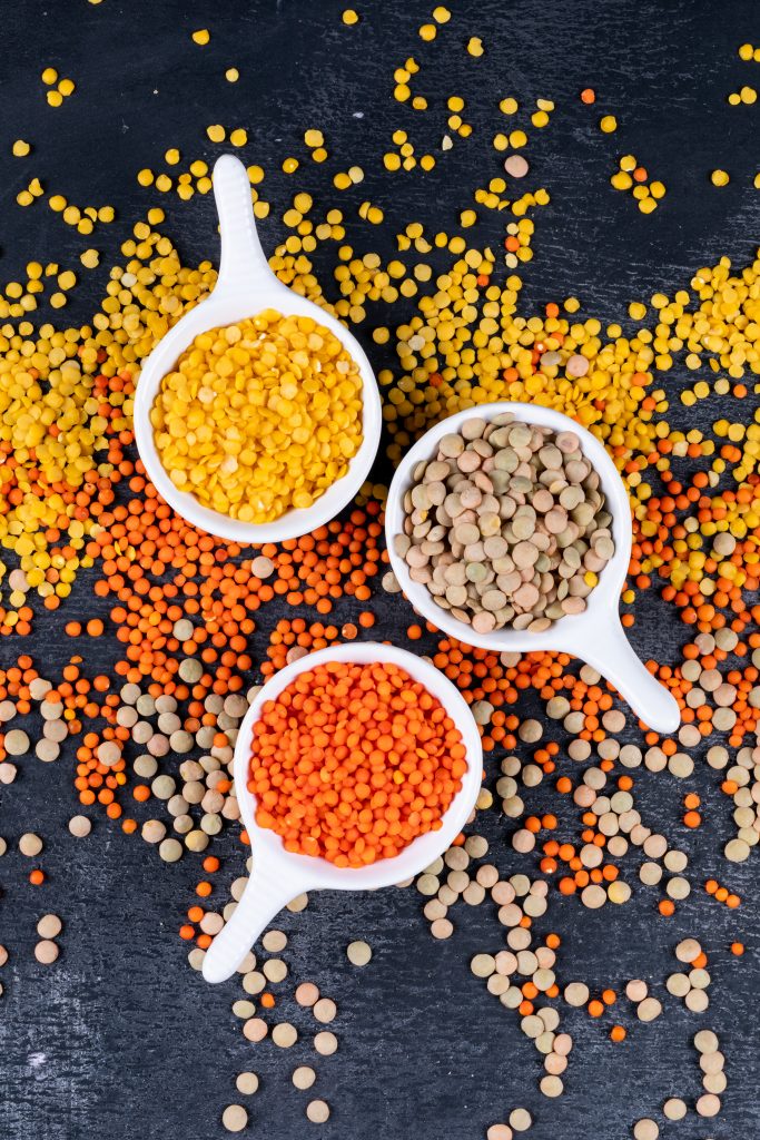 A top view of different lentils in mini white spice bowls arranged on a black stone background.