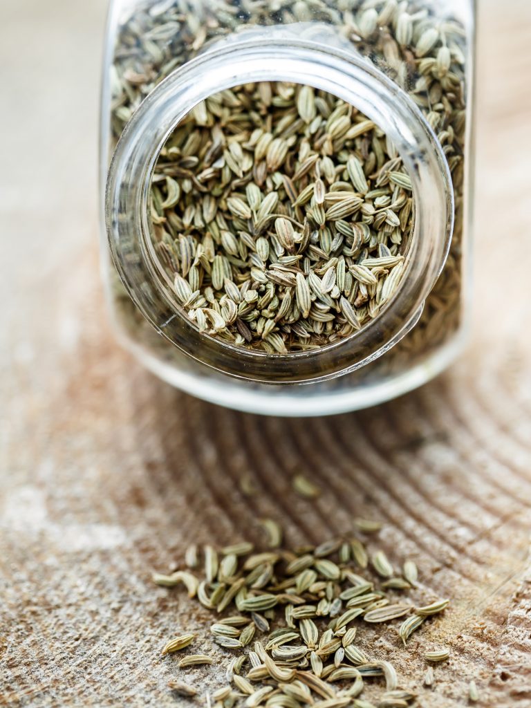 An aesthetic shot of dry fennel seeds neatly placed in a glass jar, with a few scattered around it on a clean surface.