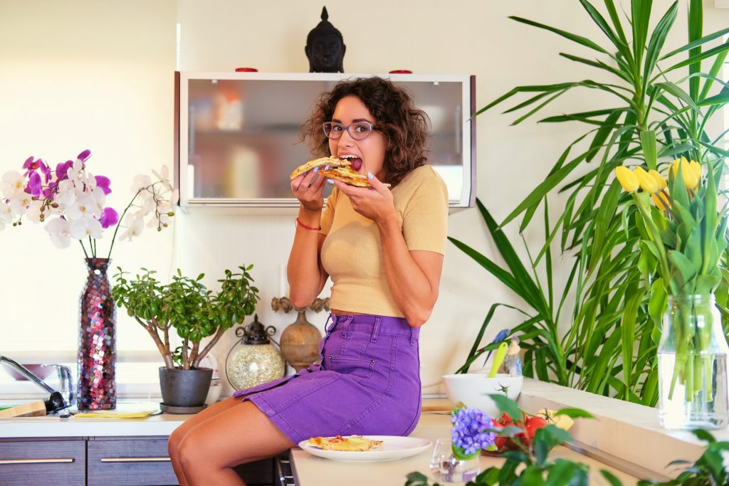 A woman eating pizza slices at a table surrounded by indoor plants, showing how food cravings can challenge dieters.