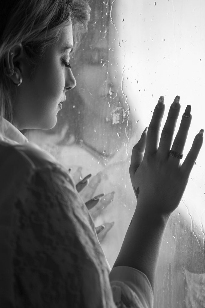 A woman appearing sad with closed eyes, lost in deep thought, standing near a window.