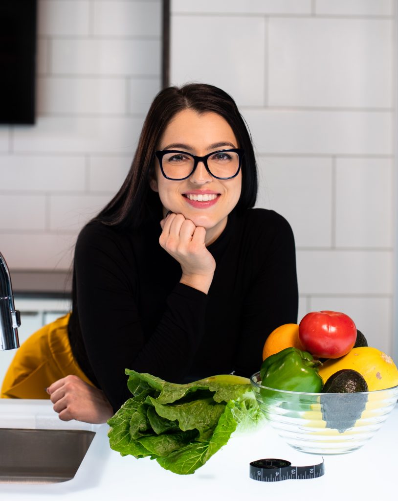 A woman candidly posing with a happy expression, with fresh vegetables neatly arranged on a white kitchen counter.