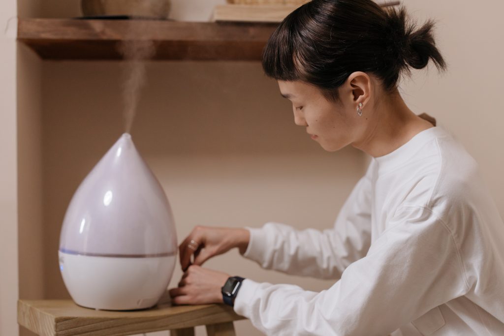 A humidifier releasing steam, with a woman sitting nearby, illustrating how the moisture helps to ease breathing.