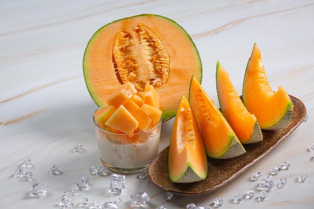 Cantaloupe cut in half, as cubes and as slices aesthetically arranged on a white surface.