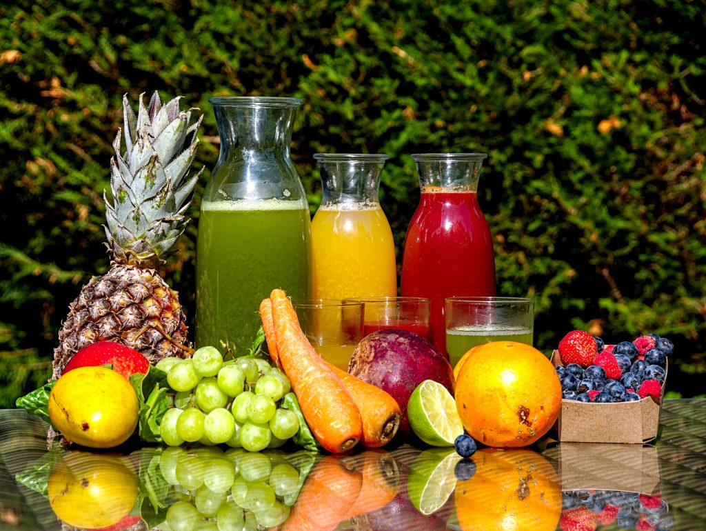 Colorful fruit juices filled in glass jars and glasses, surrounded by a variety of fresh fruits, elegantly displayed on a glass table outdoors.