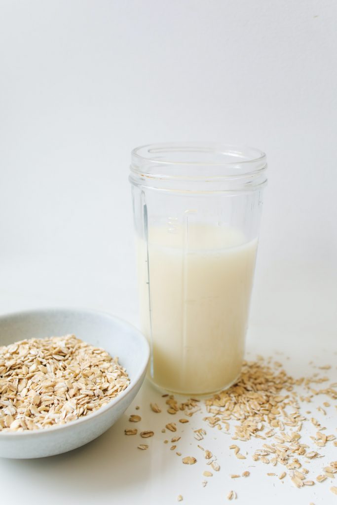 A glass of oat milk with scattered oats and a bowl of oats on a white background.