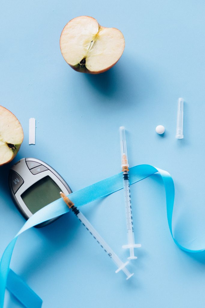 A cut apple, blood sugar monitor, insulin syringe, and medicine arranged against a blue background, symbolizing the importance of managing diabetes.