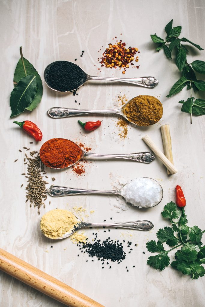 Spices like cumin, sesame seeds, coriander, turmeric, and others neatly arranged in spoons on a white surface, showcasing their vibrant colors and health benefits.