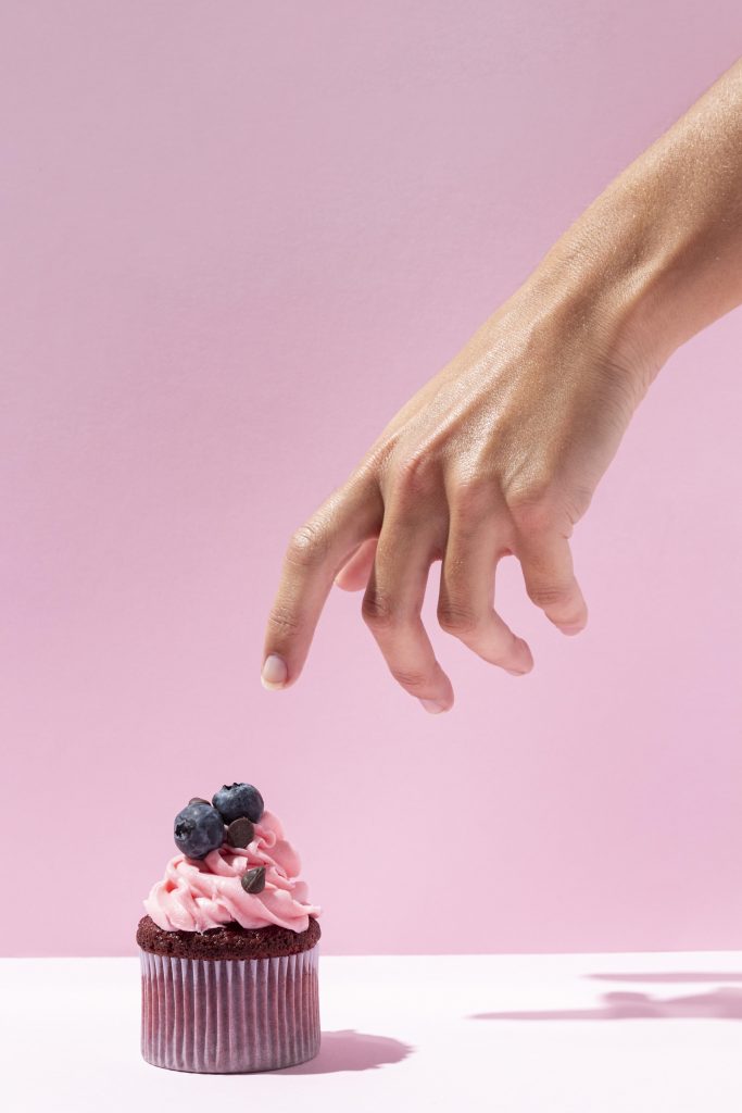 An aesthetic shot of a strawberry cupcake against a pink backdrop, highlighting the importance of indulging in your favorite foods in small portions occasionally.