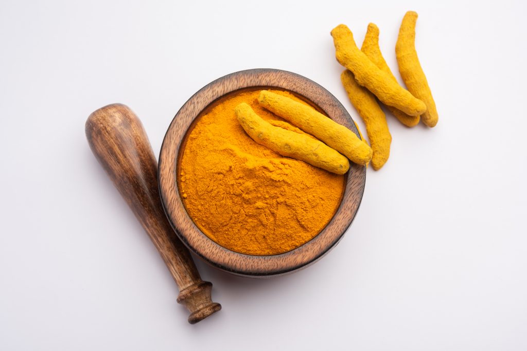 Dry turmeric dust, an antioxidant, in a mortar with a pestle, with whole turmeric roots lying nearby.