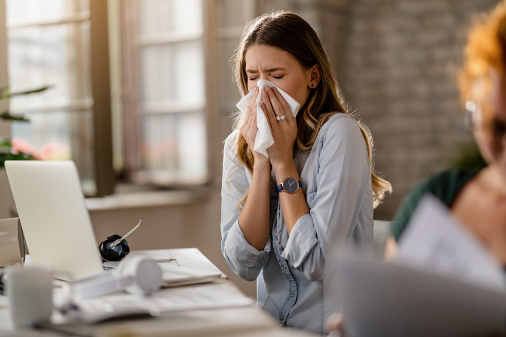 A woman at her office, sneezing, seemingly due to an allergy.