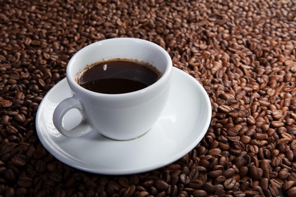 A white cup filled with coffee with a matching saucer, placed on a heap of coffee beans