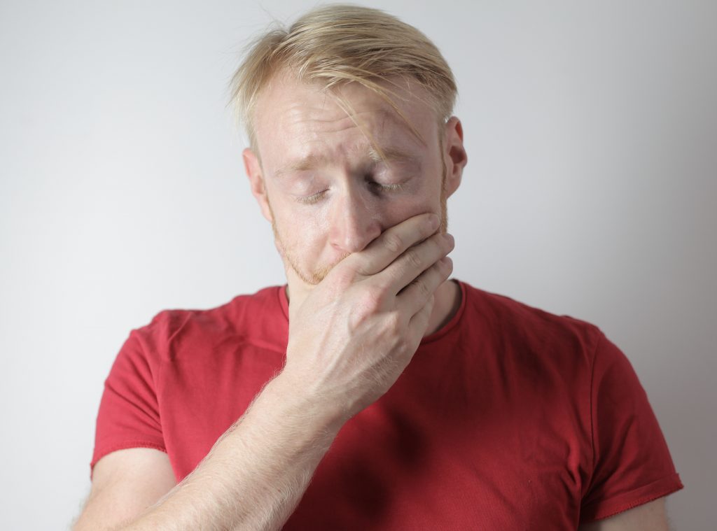 A man in a red shirt covering his face, reflecting the discomfort of mouth ulcers linked to B12 deficiency and anemia.