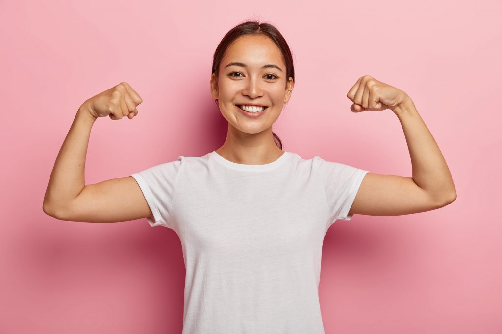 Smiling woman in a white t-shirt flexing her arms, displaying strength and positivity to boost natural defenses.