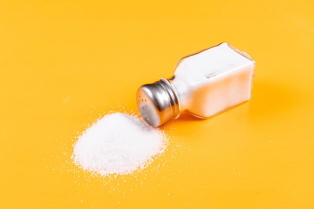 Spilled salt from a tipped-over shaker on a yellow background, symbolizing the need to reduce salt intake for hypertension control.