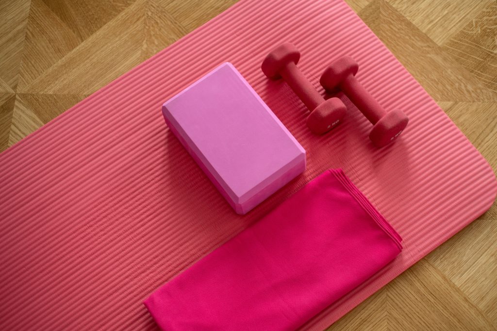 A pink yoga mat with a block, dumbbells, and a towel on a wooden floor, ideal for easing period cramps.