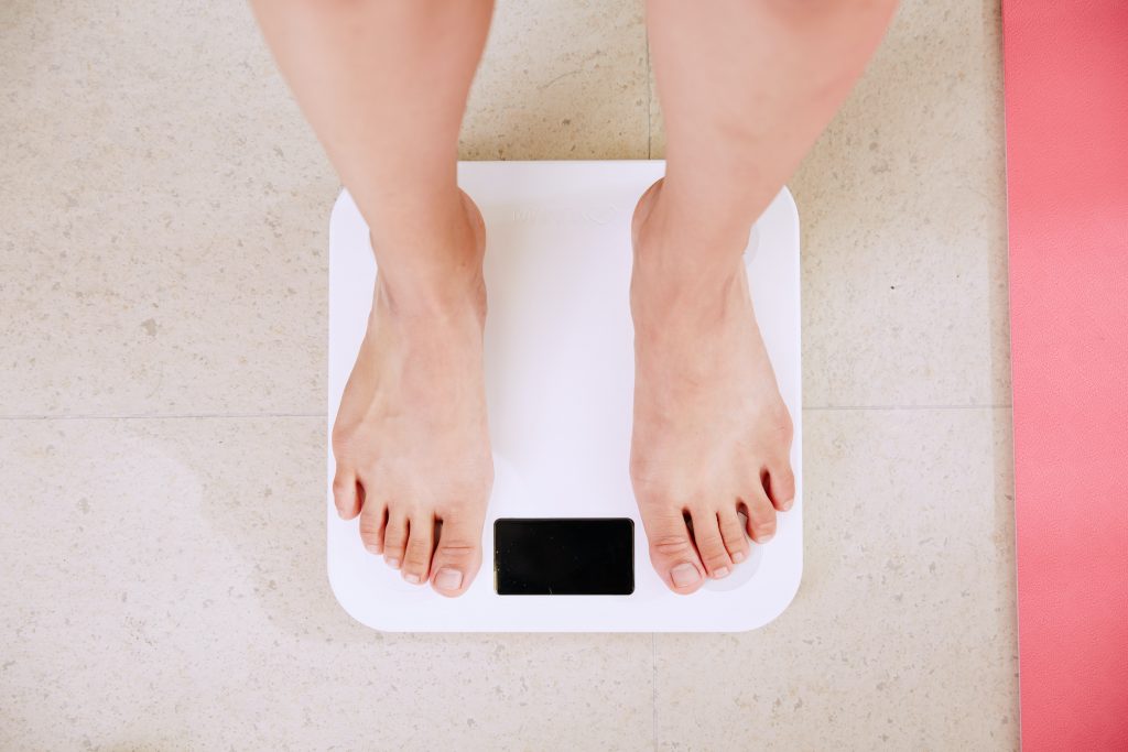 Bare feet on a weight scale on a tiled floor, next to a pink mat, emphasizing the importance of maintaining a healthy weight.