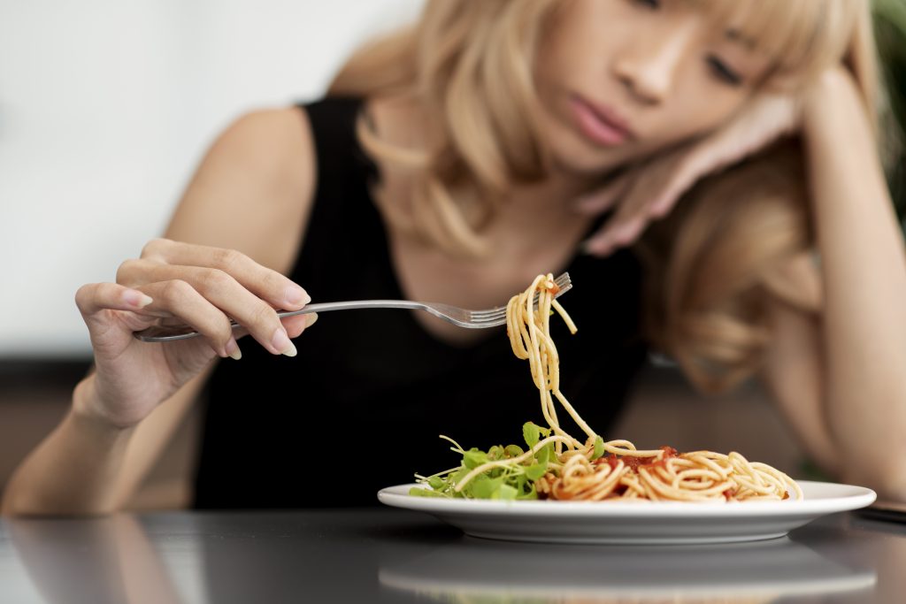 Stressed girl eating noodles with a fork, craving quick comfort with high-calorie food.