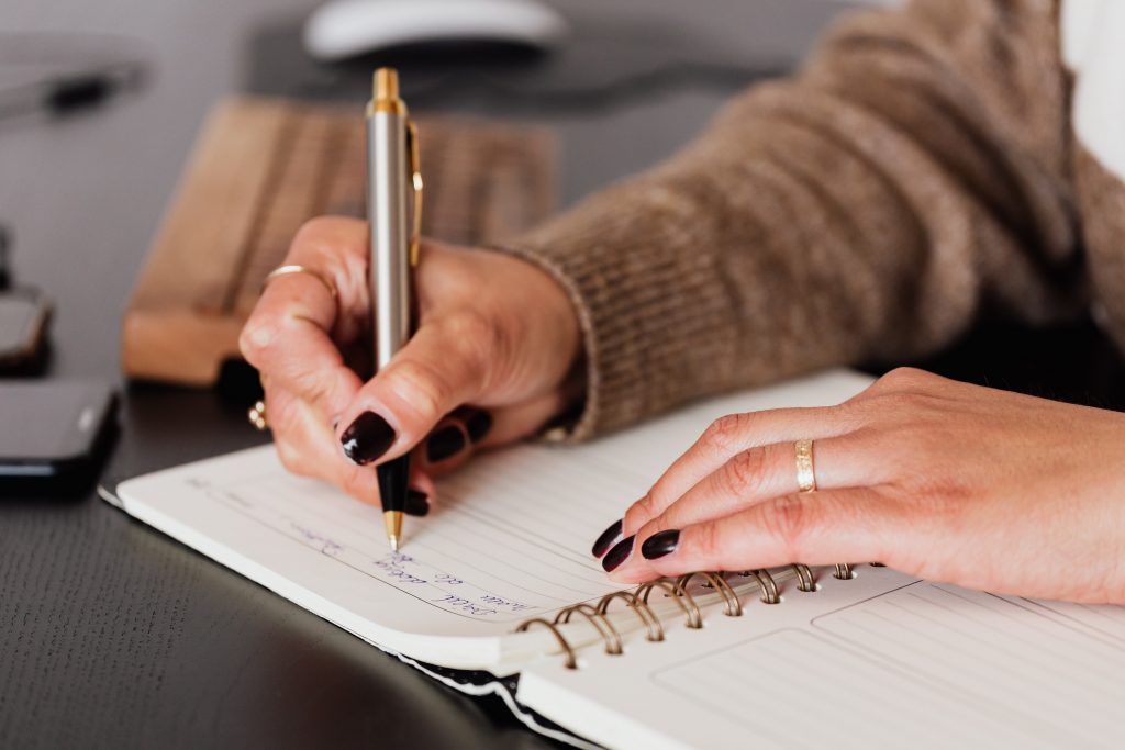 A person with black nail polish writing in a notebook with a gold pen, highlighting how tracking supports mindful eating.