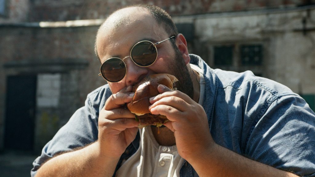 A man in sunglasses and a blue shirt eating a burger outdoors, highlighting challenges in weight loss and ways to overcome them.