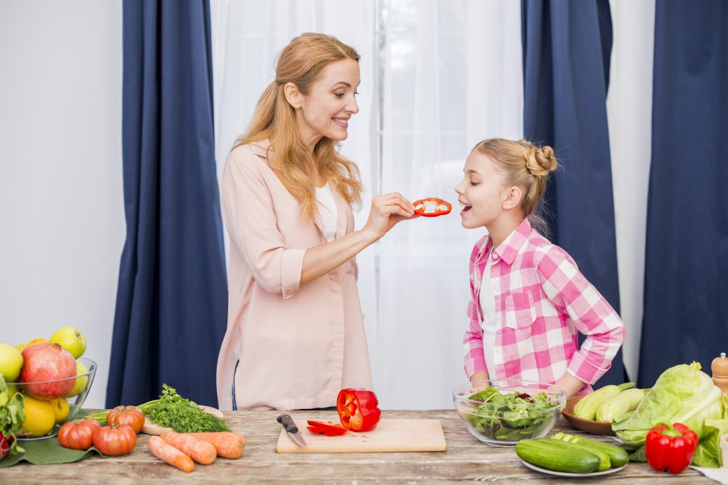 A mother feeding her daughter a slice of red bell pepper, emphasizing the importance of vegetables to avoid health problems.