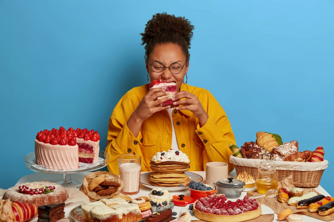 A cheerful woman with a red cake slice amidst desserts, illustrating the impact of stress on eating habits and weight gain.