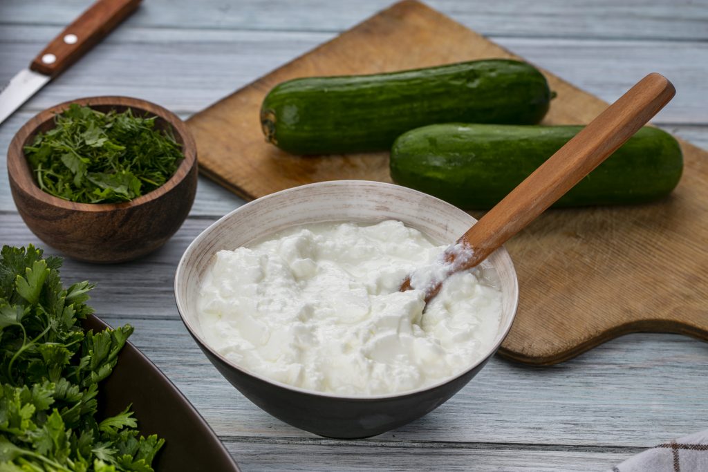 Yogurt bowl with spoon, cucumbers on a cutting board, and cilantro, packed with potassium, calcium, and probiotic benefits.