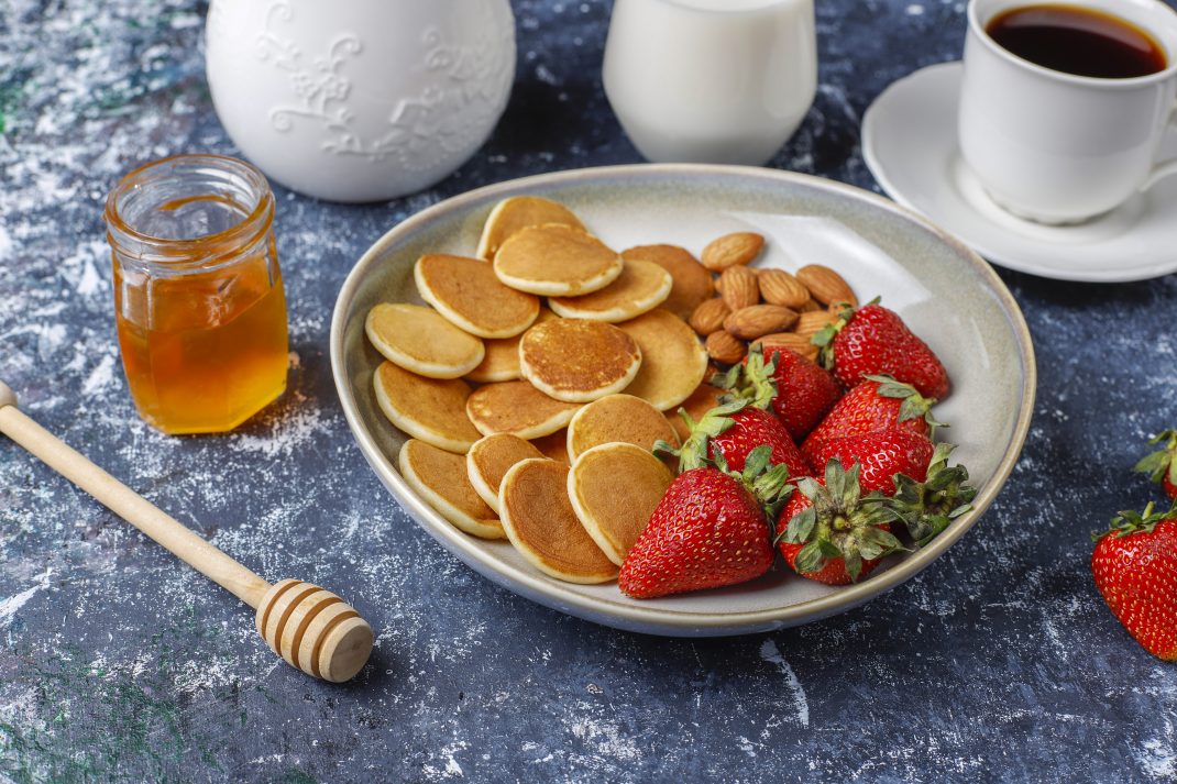 A plate of mini pancakes with strawberries, almonds, honey, and coffee, ideal for enjoying either raw or pasteurized honey.