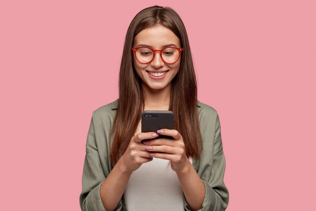 A happy young girl wearing red spectacles, texting on her phone, reminding to take breaks and use speech-to-text to avoid joint pain.