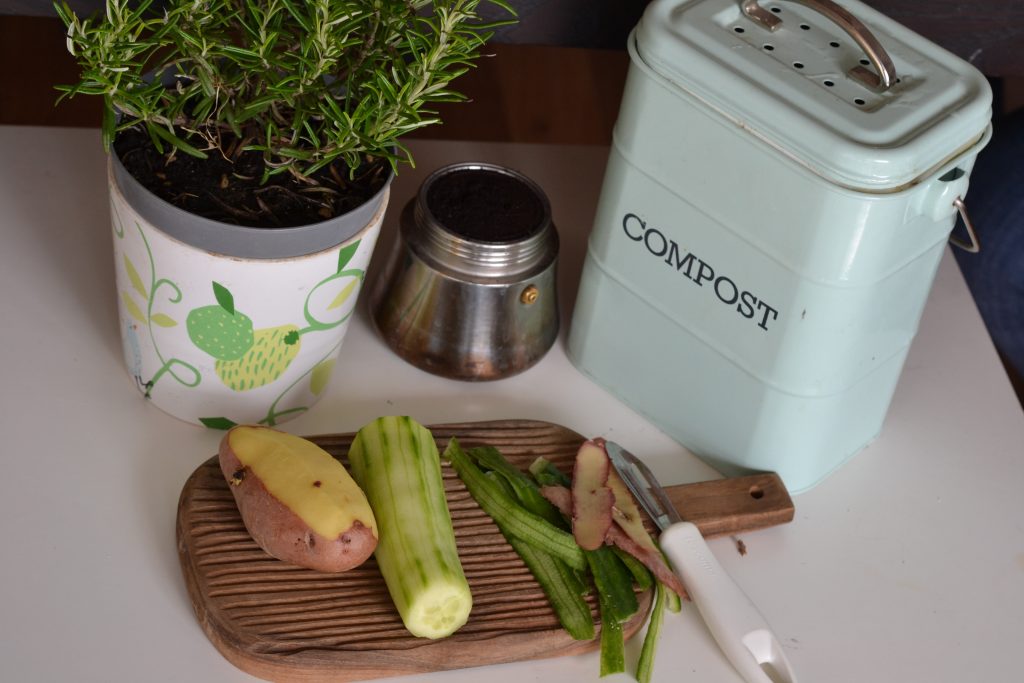 A countertop compost bin placed alongside vegetable leftovers, and an indoor herb plant.