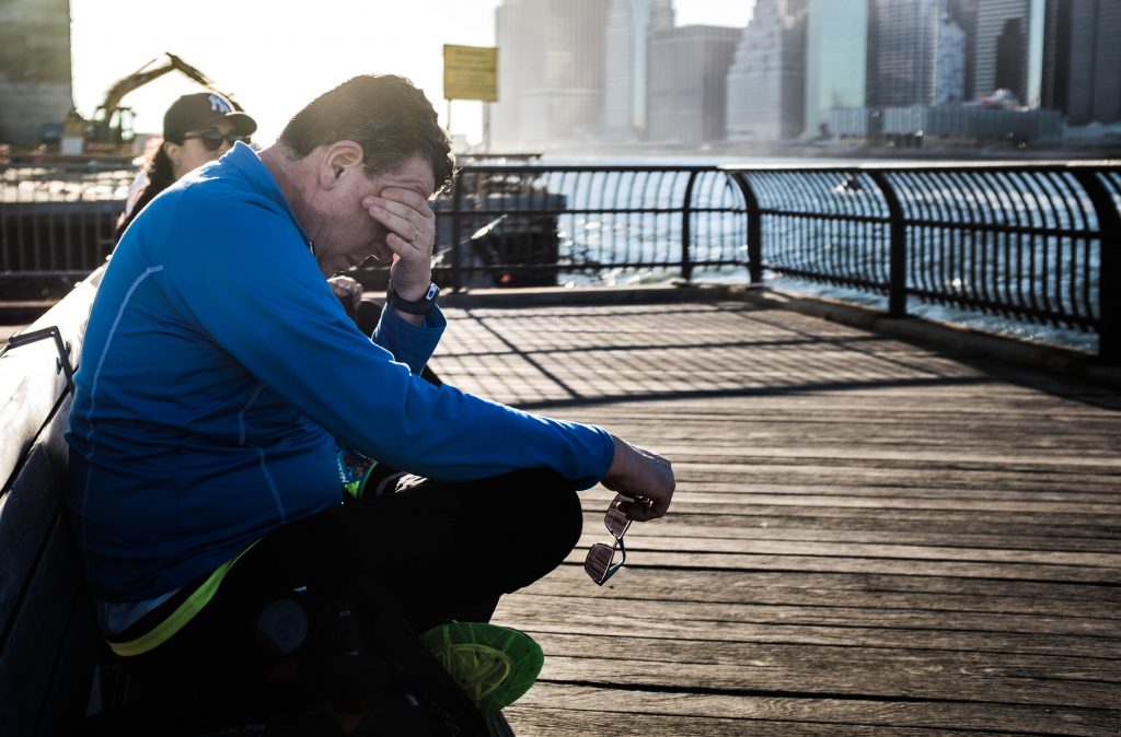 A stressed man in a blue jacket sitting on a bench, holding glasses, with his head in his hand, reflecting fatigue.