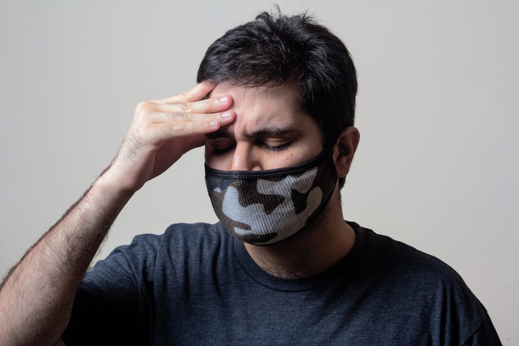 A man wearing a mask, holding his forehead with a distressed expression, visibly suffering from a severe headache.