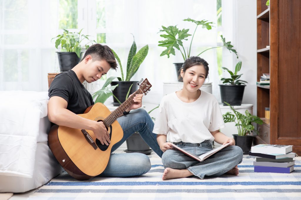 Two friends sitting on the floor in a well-ventilated room, with the boy playing guitar and the girl studying.