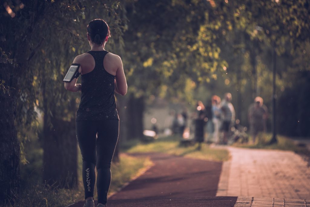 A woman jogging in a park with her phone strapped to her forearm, highlighting how excercising can reduce bloating.