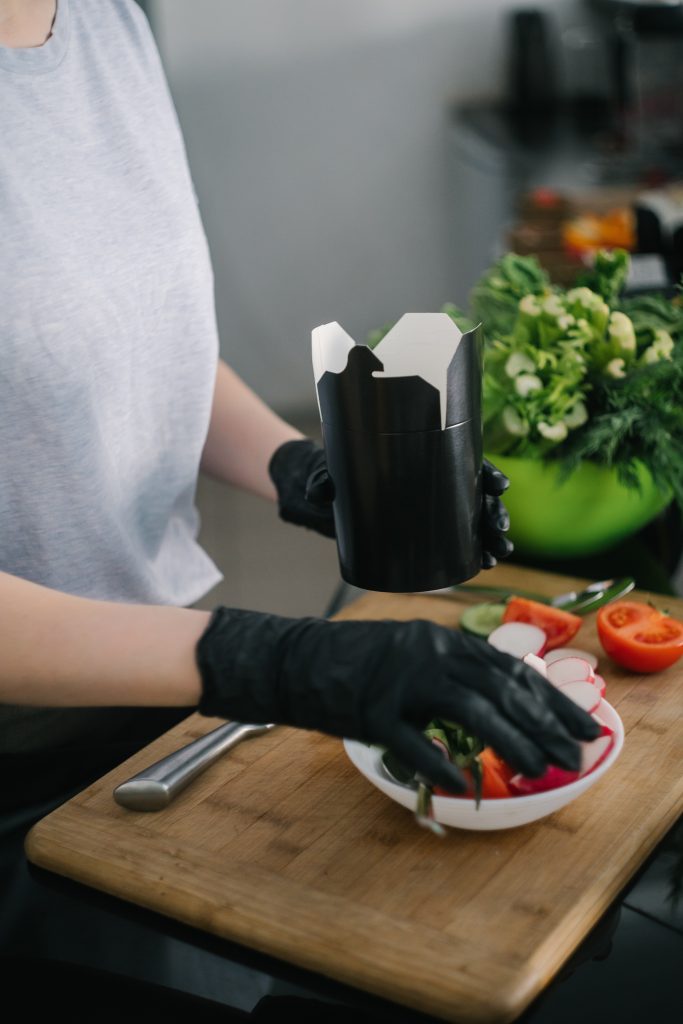 A person in black gloves prepares and packs a fresh vegetable salad on a bowl, promoting portion control to prevent overeating.