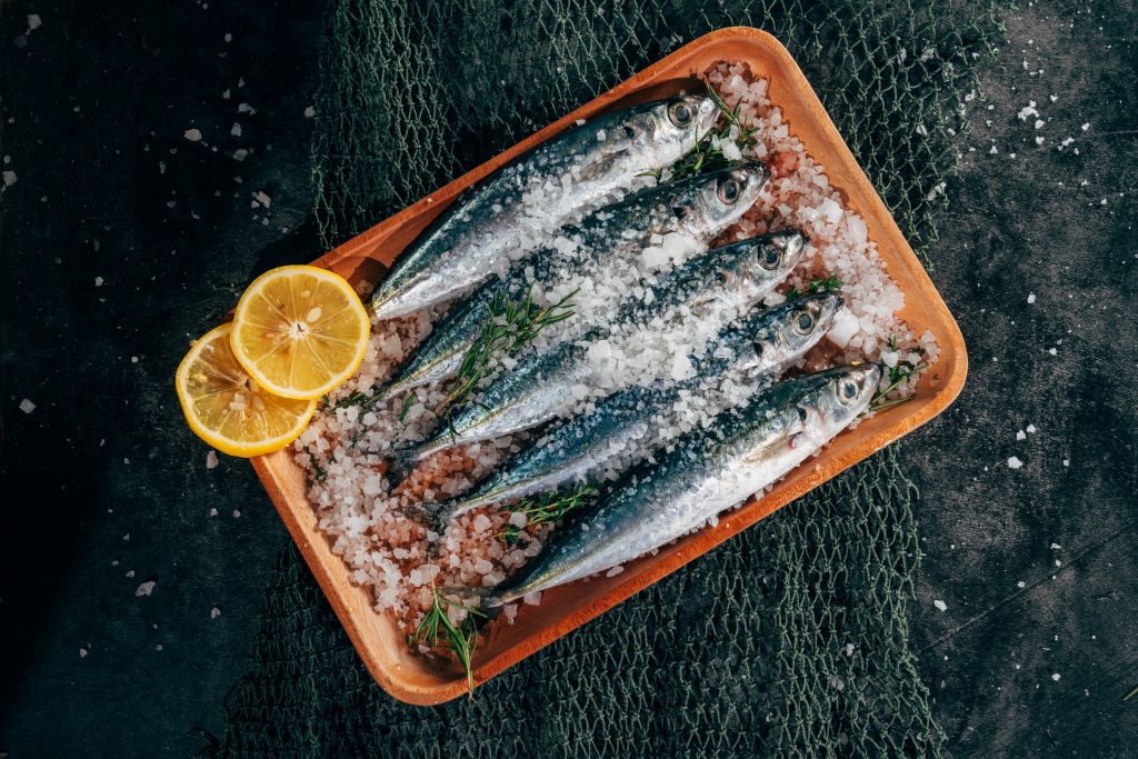 Sardines garnished with rock salt, herbs, and lemon, neatly arranged in a small tray.