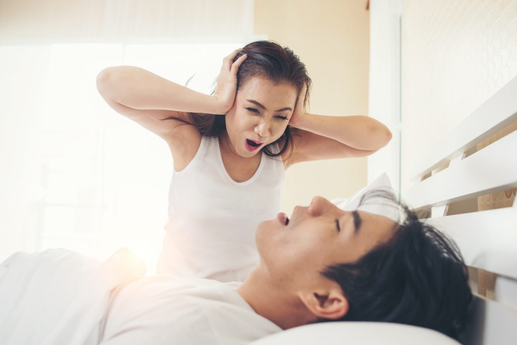 A sleep-deprived woman screaming at her partner, who is deep asleep and snoring.