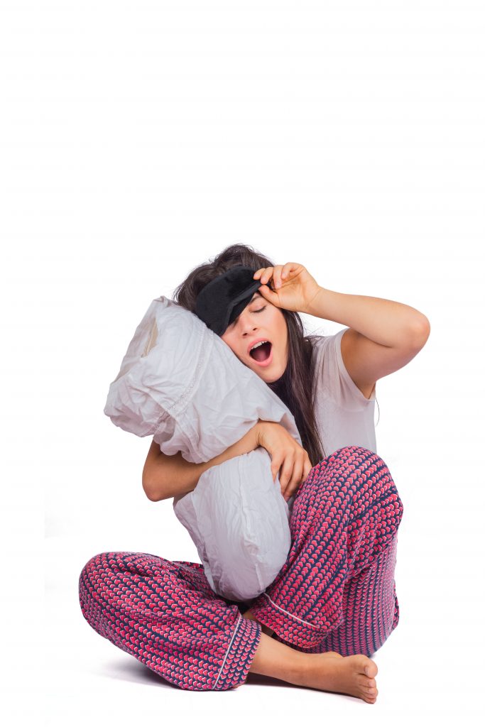 A woman sitting with a pillow, yawning and struggling to keep her eyes open, clearly feeling sleepy.