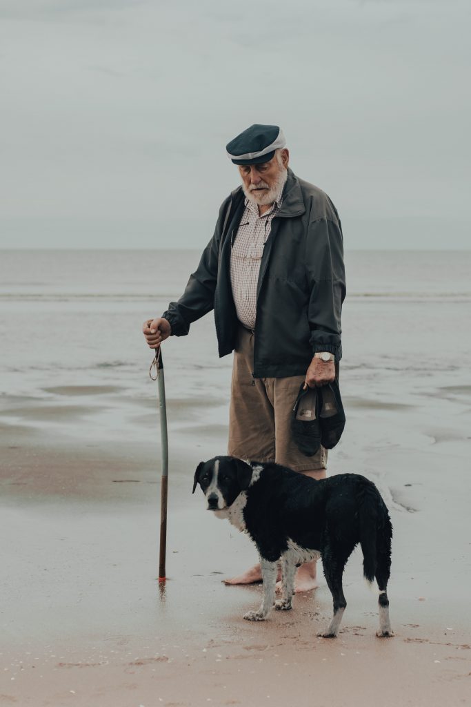 An elderly man walking along the beach with his pet dog.