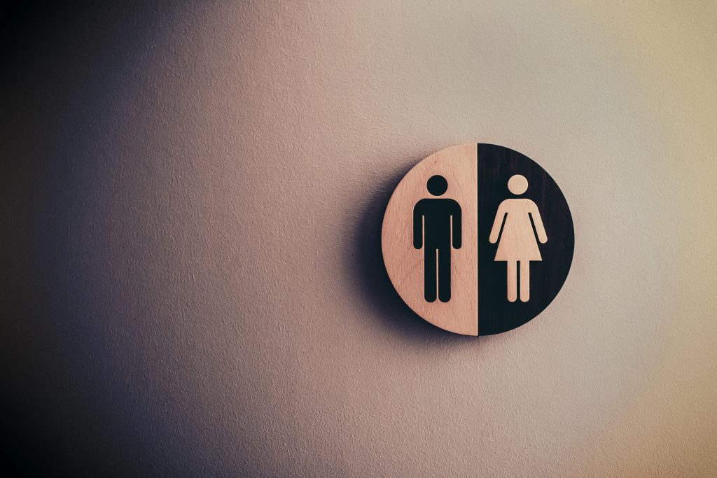 A wooden unisex restroom sign with male and female symbols on a textured wall, offering tips for frequent washroom visits.