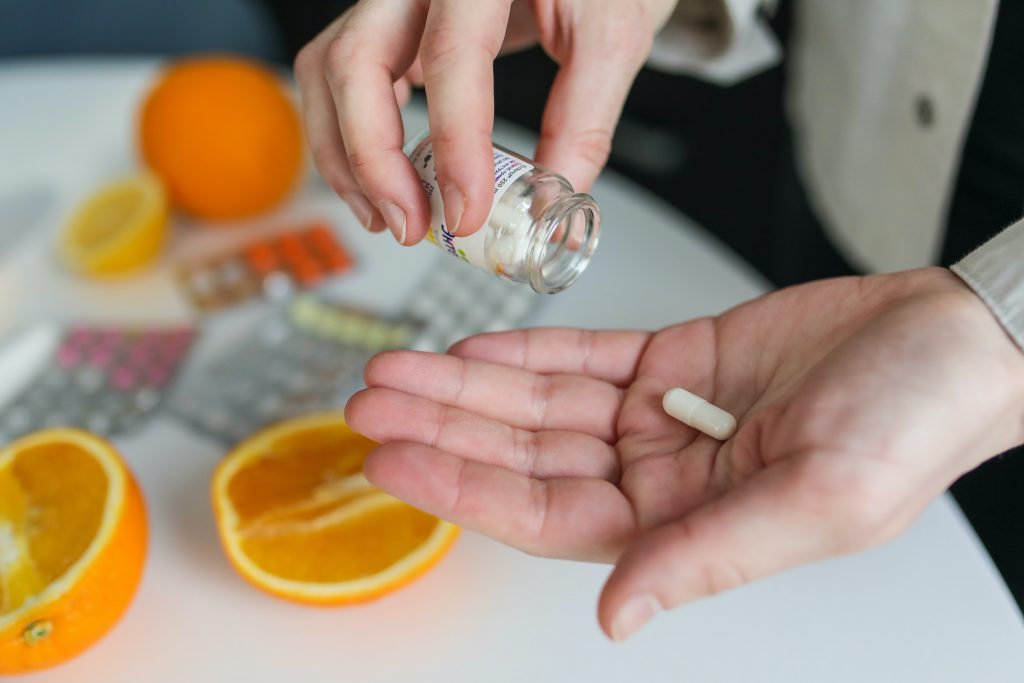 A person pouring a vitamin pill, with oranges and water on the table, symbolizing vitamin C’s help in absorbing iron.