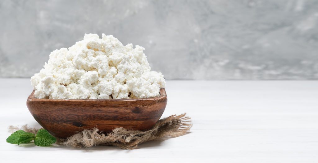 Wooden bowl with white cottage cheese on a white surface, garnished with a green leaf. A low-fat, low-calorie dairy option.