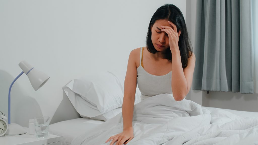 A young woman sitting on a bed, holding her head with a pained expression, possibly experiencing a headache or illness.